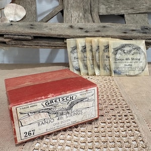 May include: A vintage red Gretsch banjo strings box with a label that reads "Banjo 4th Strings". Several individual string packets are behind the box. The box and packets are on a textured surface. The box is marked "267".