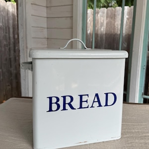 May include: A white metal bread box with a handle on the lid and a side handle. The word "BREAD" is printed in navy blue on the front. The bread box is rectangular and has a slightly distressed appearance.