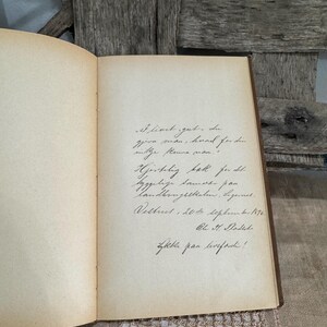 May include: An open antique book with handwritten script on aged, cream-colored pages. The book's binding is visible, and the pages show signs of age. The background includes weathered wood.