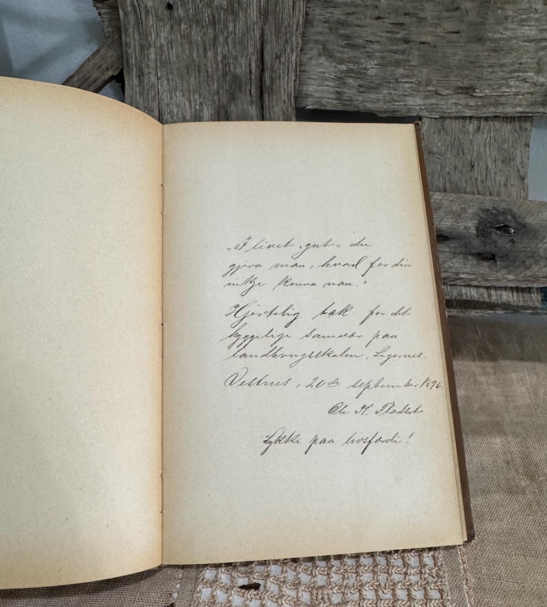 May include: An open antique book with handwritten script on aged, cream-colored pages. The book's binding is visible, and the pages show signs of age. The background includes weathered wood.