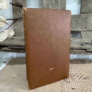 May include: A vintage hardcover book with a worn, brown leather cover. The book is slightly angled, revealing the aged edges and spine. The background features a rustic wooden surface and decorative elements.