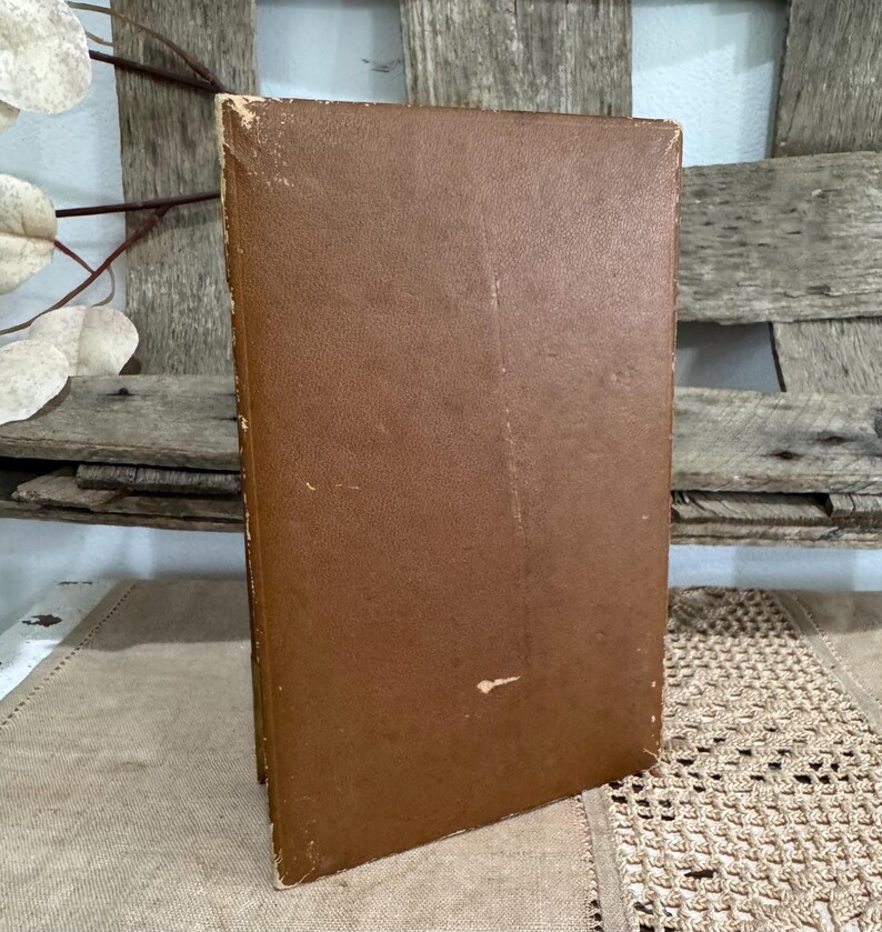 May include: A vintage hardcover book with a worn, brown leather cover. The book is slightly angled, revealing the aged edges and spine. The background features a rustic wooden surface and decorative elements.