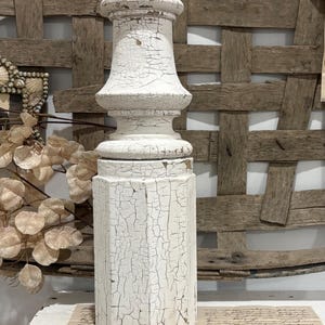 May include: A distressed white wooden pillar with a decorative, aged appearance. The pillar has a textured, cracked paint finish, and a classic, turned design. The base is octagonal, and the top has a rounded, tiered shape. This vintage-style pillar is suitable for home decor.