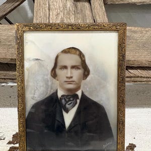 May include: A vintage portrait of a person with light brown hair, wearing a dark suit jacket and a dark cravat, encased in a rectangular gold-toned metal frame with an embossed scrollwork pattern along the border and a signature in the corner.