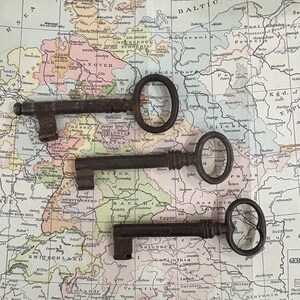 May include: Three antique iron skeleton keys with oval bows and cylindrical shafts, each featuring a distinct bit pattern at the end. The keys are dark gray and brown in color, showing signs of oxidation and age, arranged horizontally on a map.