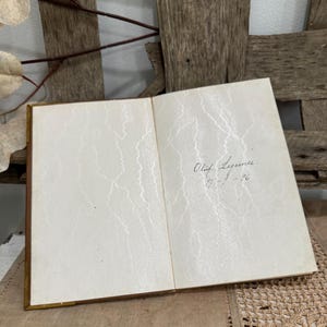 May include: An open book with aged, cream-colored pages. The pages have a subtle, textured pattern. Handwritten text in black ink is visible on the right page, including a name and a date. The book has a gold-colored spine.