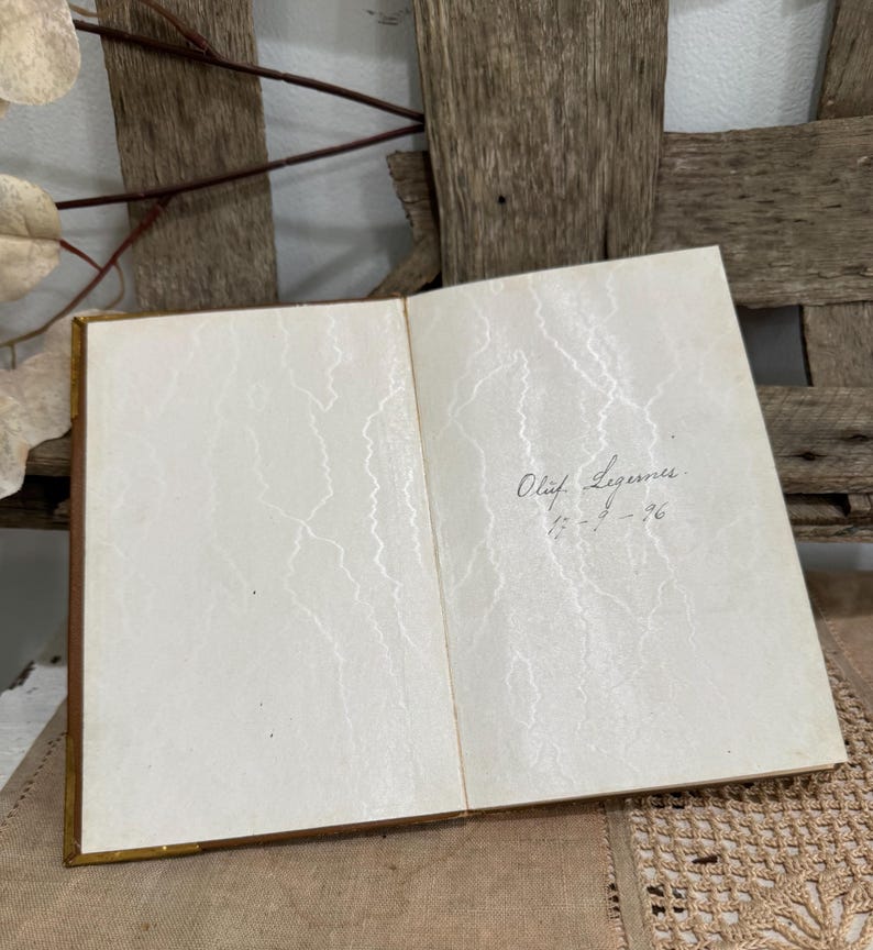 May include: An open book with aged, cream-colored pages. The pages have a subtle, textured pattern. Handwritten text in black ink is visible on the right page, including a name and a date. The book has a gold-colored spine.