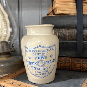 May include: A beige stoneware cream jar with a blue printed label that reads Carricks Cumberland Dairy Co Ltd Pure Thick Cream Fresh Daily Low Row Nr Carlisle. The jar has a wide mouth and a tapered body with a smooth, glazed finish.