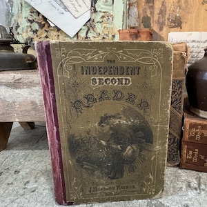 May include: An antique book titled "The Independent Second Reader" with a faded cover featuring an illustration of a woman. The book has a worn, aged appearance with a maroon spine and visible wear on the edges, suggesting its age and use.