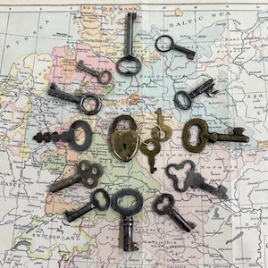 May include: A collection of various antique metal keys and a single heart-shaped padlock arranged on a vintage map of Europe. The keys vary in size, shape, and metal finish, including dark iron, brass, and steel tones, scattered across the paper.