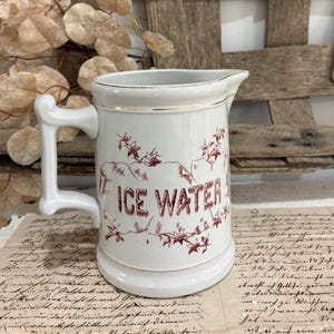 May include: A white ceramic pitcher with a gold rim and a handle. The front features the text "ICE WATER" in a dark red, decorative font, surrounded by a brown, hand-drawn illustration of ivy leaves and vines wrapping around the body.