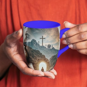 May include: A blue-handled coffee mug with a blue interior, depicting a landscape scene with a cross, figures, and mountains. The mug is held by a person wearing a white top and a red overall dress.