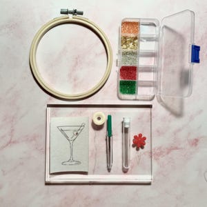 Könnte beinhalten: Ein Stickset mit einem Holzrahmen, einem transparenten Behälter mit bunten Perlen, einer Leinwand mit Martini-Glas-Design, Faden, einem Stift, einer Nadel und einer roten Blumen-Anstecknadel.
