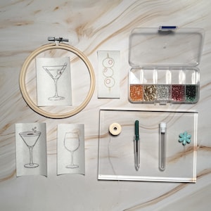 May include: A collection of embroidery supplies and tools, including a wooden hoop with a martini design, glitter, needles, and a clear plastic organizer. The image also features embroidered cocktail glasses and olive designs.