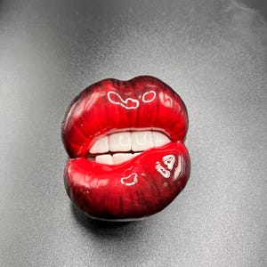 Puede incluir: Una escultura roja brillante de un par de labios con dientes blancos. Los labios están ligeramente entreabiertos, revelando los dientes. La escultura tiene una superficie brillante y reflectante, y el color es un rojo intenso y vibrante.
