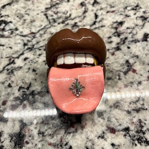 May include: A novelty sculpture of a mouth with brown lips, white teeth, and a pink tongue. The tongue has a gem in the center. The teeth have gold accents.