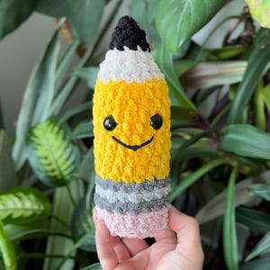 May include: A handmade, plush pencil with a smiling face. The pencil features a black tip, white eraser, yellow body, and gray and pink accents. The pencil is held in front of a background of green leaves.