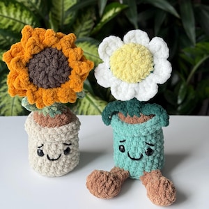 May include: Two crocheted flower pot dolls. One is a sunflower with orange petals and a brown center, the other is a daisy with white petals and a yellow center. Both have smiling faces and are sitting on a white surface.