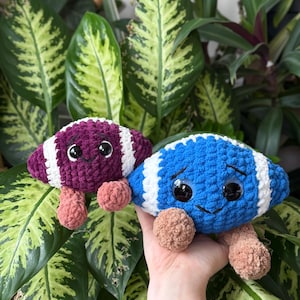 Puede incluir: Dos juguetes de ganchillo con forma de balón de fútbol. Uno es morado con rayas blancas, el otro es azul con rayas blancas. Ambos tienen ojos saltones negros grandes y pies de color canela. Los juguetes están colocados sobre un fondo de follaje verde.