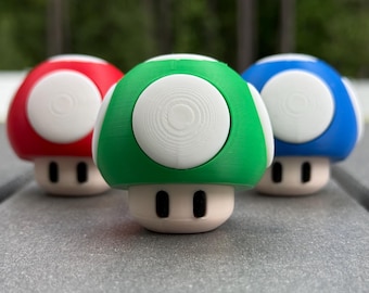May include: Three colorful mushroom-shaped figurines in red, green, and blue, each with a white circular top and a beige base. The mushrooms have two black oval-shaped eyes. The figurines are arranged in a row on a gray surface.