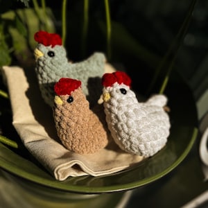 May include: Three crocheted chicken figurines in shades of brown, green, and white, each with a red comb and yellow beak. The chickens are arranged on a beige cloth inside a green bowl, with a blurred background of greenery.