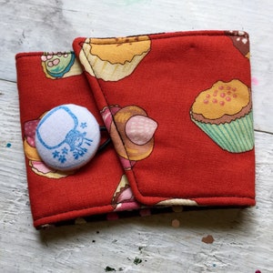 Puede incluir: Un brazalete de tela roja con un estampado de magdalenas y cierre de botón. El botón blanco tiene un diseño azul. El brazalete está diseñado para envolver un artículo, como una taza de café o una muñeca.