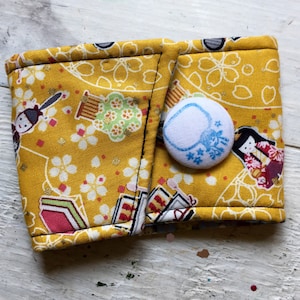 May include: A yellow fabric coffee sleeve with a Japanese-inspired pattern featuring dolls, fans, and cherry blossoms. The sleeve is secured with a white button with a blue design.