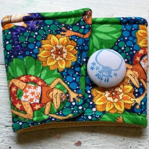 May include: A fabric coffee sleeve with a button fastening. The sleeve displays a lively pattern of frogs, flowers, and water droplets in orange, green, blue, and yellow. A small, round button with a blue design fastens the sleeve.
