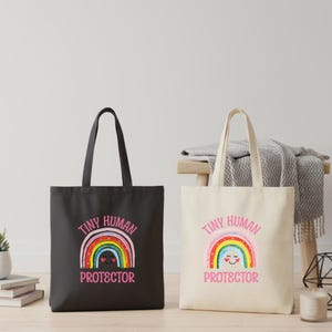 May include: Two tote bags, one black and one cream-colored, each featuring a rainbow design with the text "Tiny Human Protector" in pink. The rainbow has a smiling face. The bags have long handles.