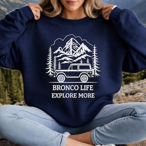 May include: Navy blue sweatshirt with a white graphic of a mountain range, trees, and a vintage SUV. The text "BRONCO LIFE EXPLORE MORE" is printed below the graphic. The sweatshirt is a crew neck style.