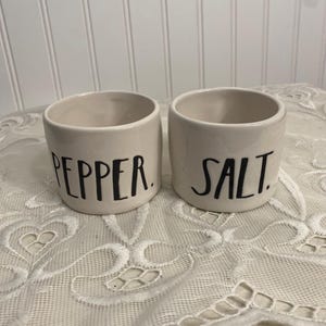 May include: Two off-white ceramic salt and pepper shakers. The word "PEPPER" is printed in black on one shaker, and "SALT" is printed on the other. The shakers are cylindrical with a wide opening and sit on a white lace tablecloth.