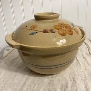 Könnte beinhalten: Beige Keramik-Auflaufform mit Deckel. Das Gericht hat ein Blumendesign in Orange, Braun und Blau. Zwei blaue Streifen umrunden die Basis. Der Deckel hat einen kleinen Griff und Seitengriffe für eine einfache Handhabung.