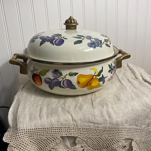 Könnte beinhalten: Cremefarbener Emaille-Topf mit Deckel, verziert mit einem Fruchtdesign mit Pflaumen, Birnen und Trauben. Der Topf hat messingfarbene Griffe und einen dekorativen Knopf. Ein Vintage-Küchenartikel.