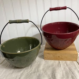 May include: Two ceramic bowls with wire handles. One is deep green, the other is burgundy. Both have a textured pattern around the outside and wooden handles. The bowls are sitting on a white cloth.