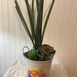 May include: A decorative metal bucket with a floral design, filled with artificial succulents and tall green grass-like stems. The bucket has a distressed finish and a small handle. The floral design features yellow, purple, and red flowers.