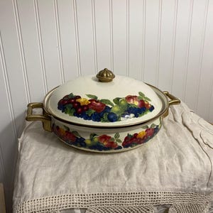 May include: Cream-coloured enamelware casserole dish with a lid, featuring a colourful fruit design around the rim and on the lid. The dish has gold-toned handles and a gold knob on the lid. The overall shape is oval.