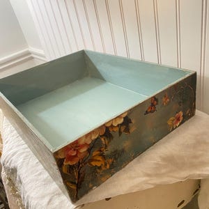 May include: A rectangular wooden tray with a light blue interior and floral artwork on the exterior. The artwork features pink and yellow flowers with butterflies. The tray is sitting on a white surface.