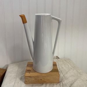 May include: A white ceramic watering can with a long, slender spout and a gold-colored tip. The handle is a simple, angular design. The watering can is displayed on a wooden block.