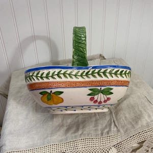 Może przedstawiać: Ceramiczny kosz z zielonym uchwytem i białą podstawą. Kosz jest ozdobiony niebiesko-zielonym wzorem liści, pomarańczowym pasem oraz ilustracjami żółtych gruszek i czerwonych wiśni. Uchwyt jest zielony.