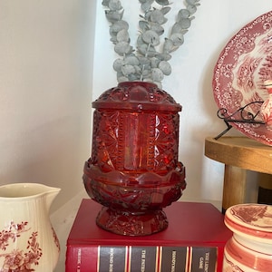 Pode incluir: Um frasco de vidro vermelho rubi, de estilo vintage, com tampa, apresentando um padrão de diamante. O frasco está em cima de um livro vermelho com letras douradas. Um jarro branco com um design floral está à esquerda, e um prato decorativo está no fundo.