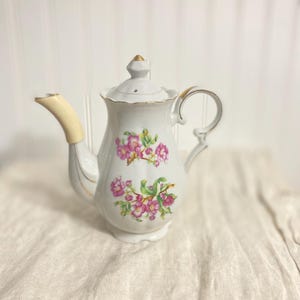 Könnte beinhalten: Weiße Porzellan-Teekanne mit einem Blumendesign aus rosa Blüten und grünen Blättern. Die Teekanne hat einen cremefarbenen Ausguss und Griff mit goldenen Zierleisten. Der Deckel ist mit einer goldenen Spitze versehen. Vintage-Teezubehör.