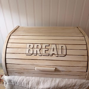 May include: A light-colored wooden bread box featuring a roll-top design with horizontal slats. The word BREAD is printed in dark, bold, block capital letters across the center of the front panel, which also includes a small, rectangular wooden handle.