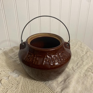 Puede incluir: Un tarro de cerámica marrón con interior oscuro y asa metálica. El tarro tiene texto en relieve alrededor del centro. El tarro está sobre un mantel de encaje color crema.