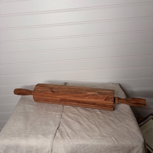 May include: A cylindrical wooden rolling pin featuring a distinct brown and tan wood grain pattern. The tool has a central barrel section and two smaller, contoured handles on each end, showcasing the natural texture and color variations of the wood.