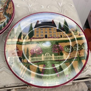 Puede incluir: Plato decorativo con un diseño de paisaje. El plato presenta una casa amarilla bajo un cielo azul, rodeada de árboles, una valla blanca y un jardín con follaje rosa y verde. El borde es de color burdeos oscuro.