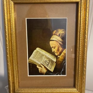 May include: A framed print of a woman reading a book. The print is in a gold-colored frame with a textured surface. The woman is wearing a head covering and a fur-lined garment. The book is open, displaying text and an illustration.