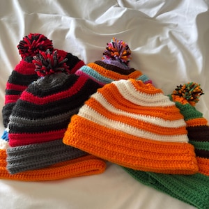 May include: A collection of colorful, handmade crochet beanies with pom-poms. The beanies feature various color combinations, including orange and white stripes, black and red stripes, and a green beanie with brown stripes. The beanies are displayed on a white surface.