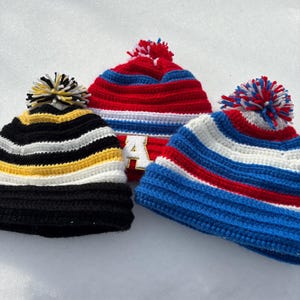 May include: Three crocheted beanies with pom-poms rest on a snowy surface. One beanie is black with yellow and white stripes, another is red with blue stripes and the letter "A", and the third is blue with red and white stripes.
