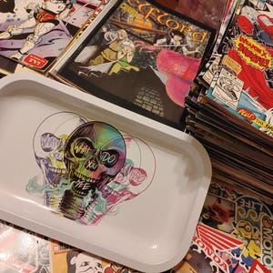 May include: A white rectangular tray with rounded corners, featuring a colorful skull design with the text "WHAT DO YOU SEE". The skull has a rainbow gradient and is surrounded by smoke-like imagery. Comic books are stacked in the background.