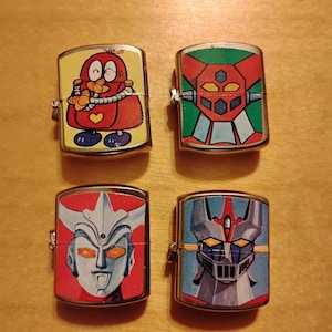 Puede incluir: Cuatro encendedores met&aacute;licos vintage con coloridos dise&ntilde;os de dibujos animados y robots. Los encendedores presentan un personaje de dibujos animados rojo, un robot con detalles en verde y rojo, un robot con fondo rojo y un robot con fondo azul.